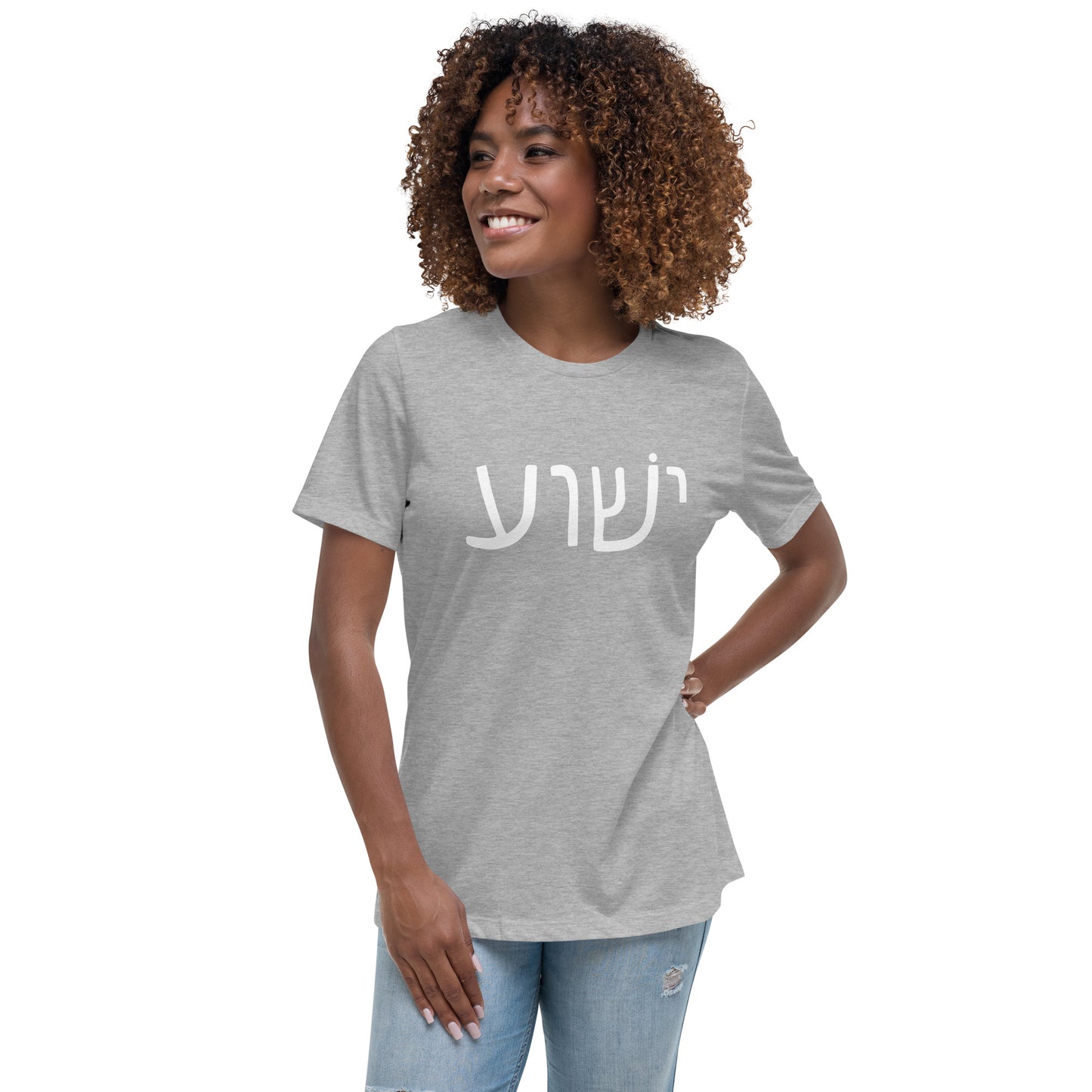A curly-haired person smiles while wearing a gray Yeshua Womens T-Shirt featuring Hebrew lettering by deusdesign, paired with light blue jeans. They stand with one hand on their hip against a plain white background.