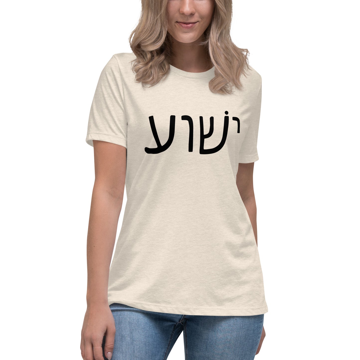 Against a plain white background, a woman with long hair wears blue jeans and the beige Yeshua Womens T-Shirt by deusdesign, featuring black Hebrew text. Her relaxed posture exudes confidence and showcases her love for unique Christian womens clothing.